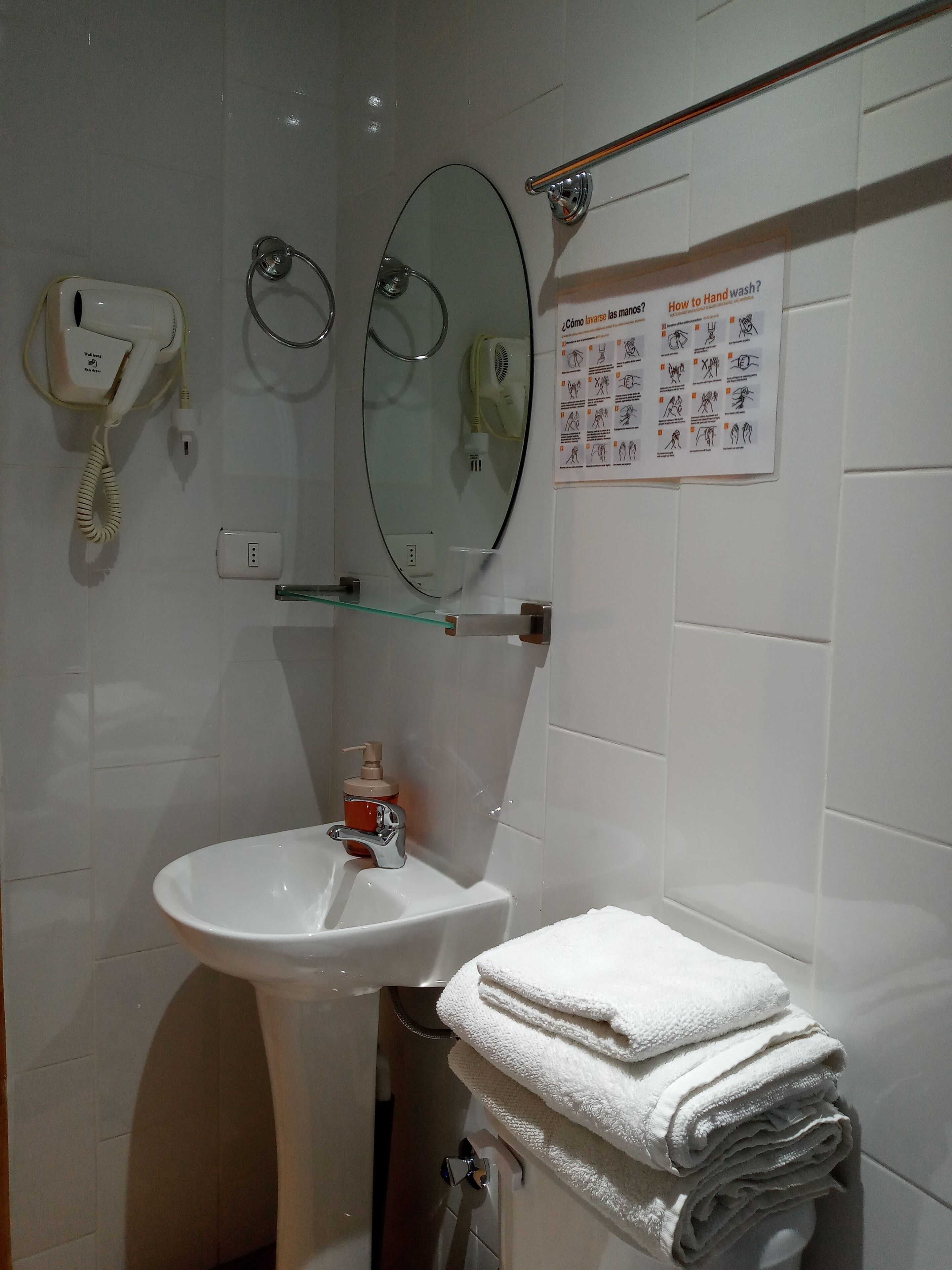 Un baño con lavamanos blanco, espejo, secador de pelo y cartel sobre lavado de manos.