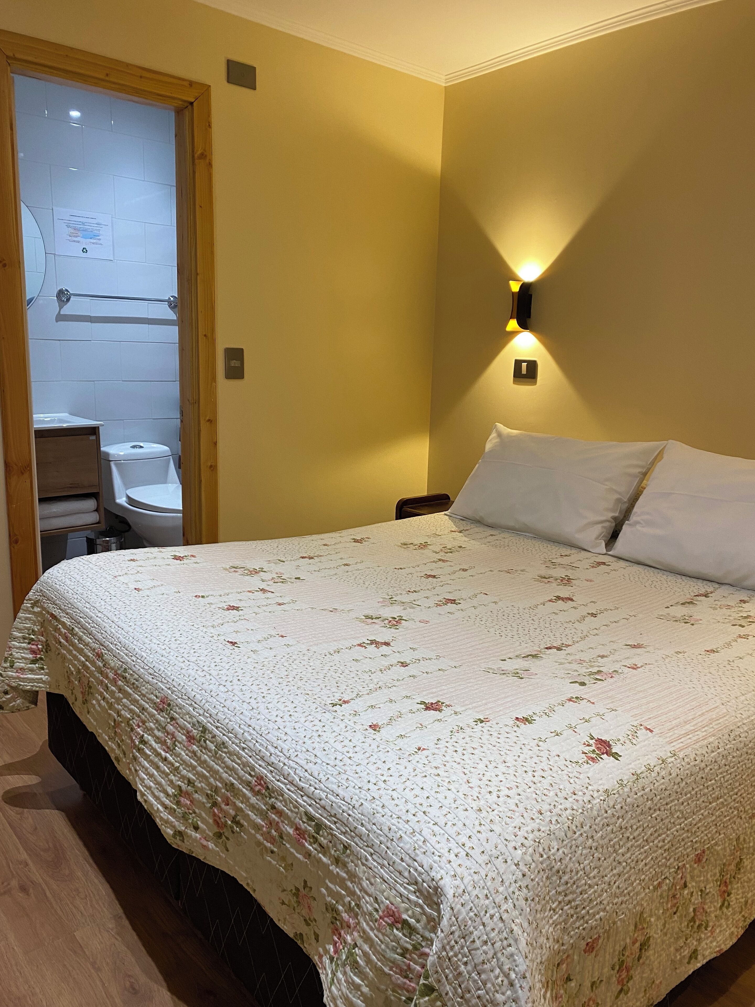 Un dormitori amb un edredó estampat amb motius florals, una porta de fusta que condueix a un bany i una làmpada fixa a la paret.