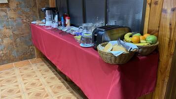 Un desayuno con mantel rojo, diferentes alimentos, un tostador y una cesta de frutas.