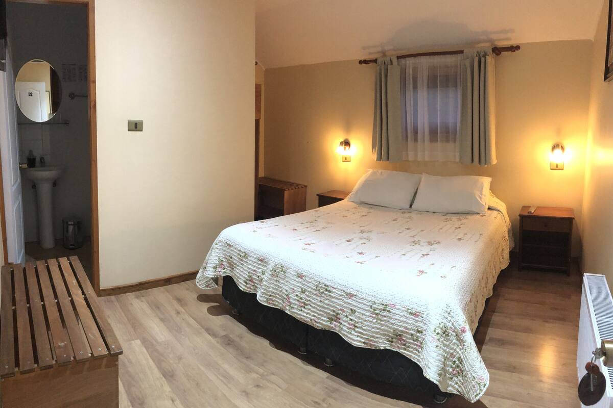 Un dormitorio con suelo de madera, una cama con colcha estampada, dos lámparas de noche, una mesita de noche, un radiador y una ventana con cortinas.