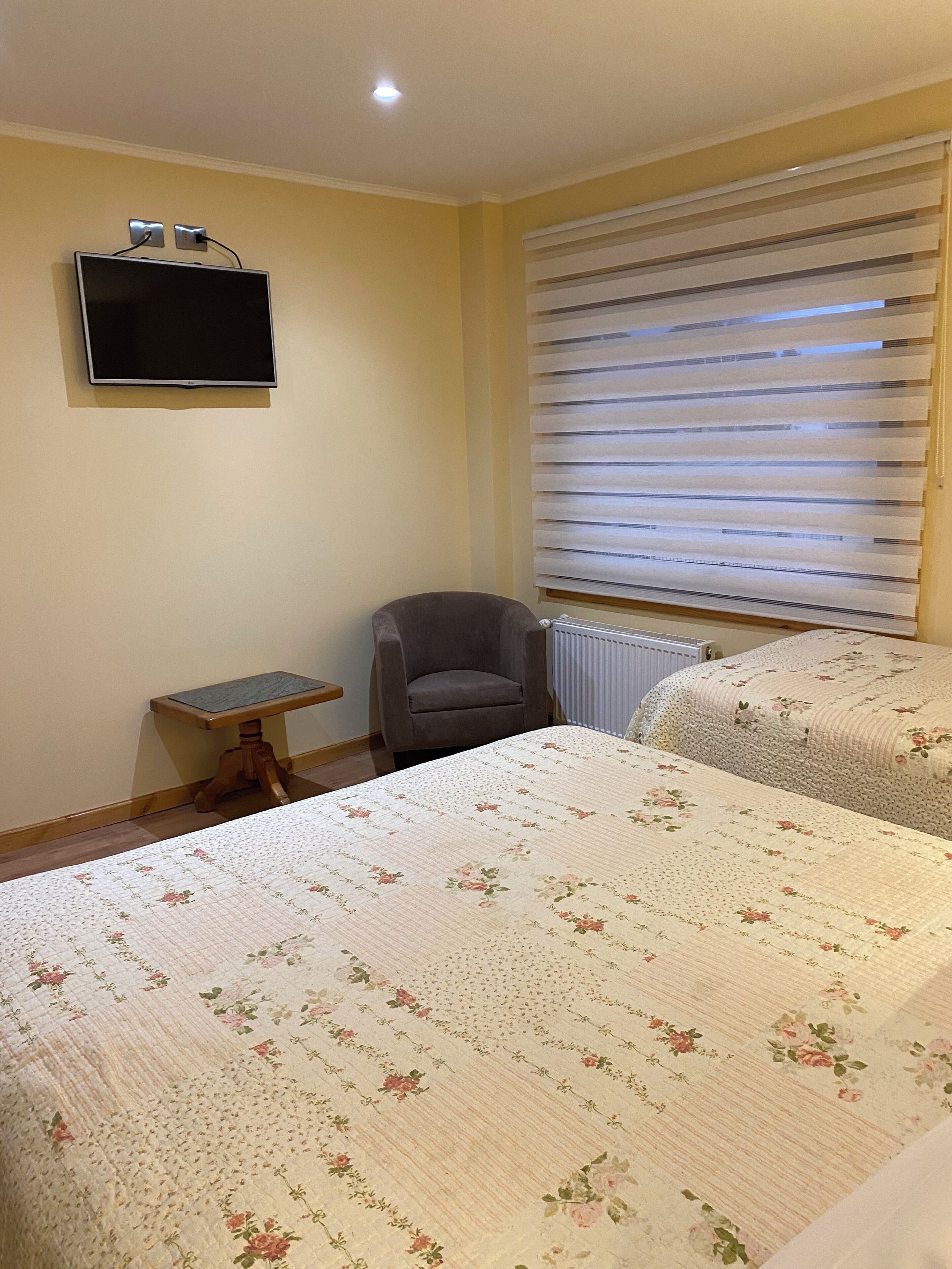 Ein Schlafzimmer mit zwei Betten, einem Sessel, einem kleinen Tisch, einem an der Wand befestigten Fernseher und einem Fenster mit Jalousien.