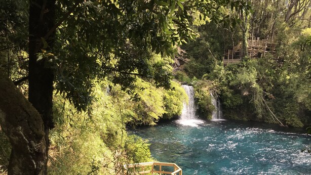 Terraza de madera con vista a un río cristalino y una cascada en un área boscosa.