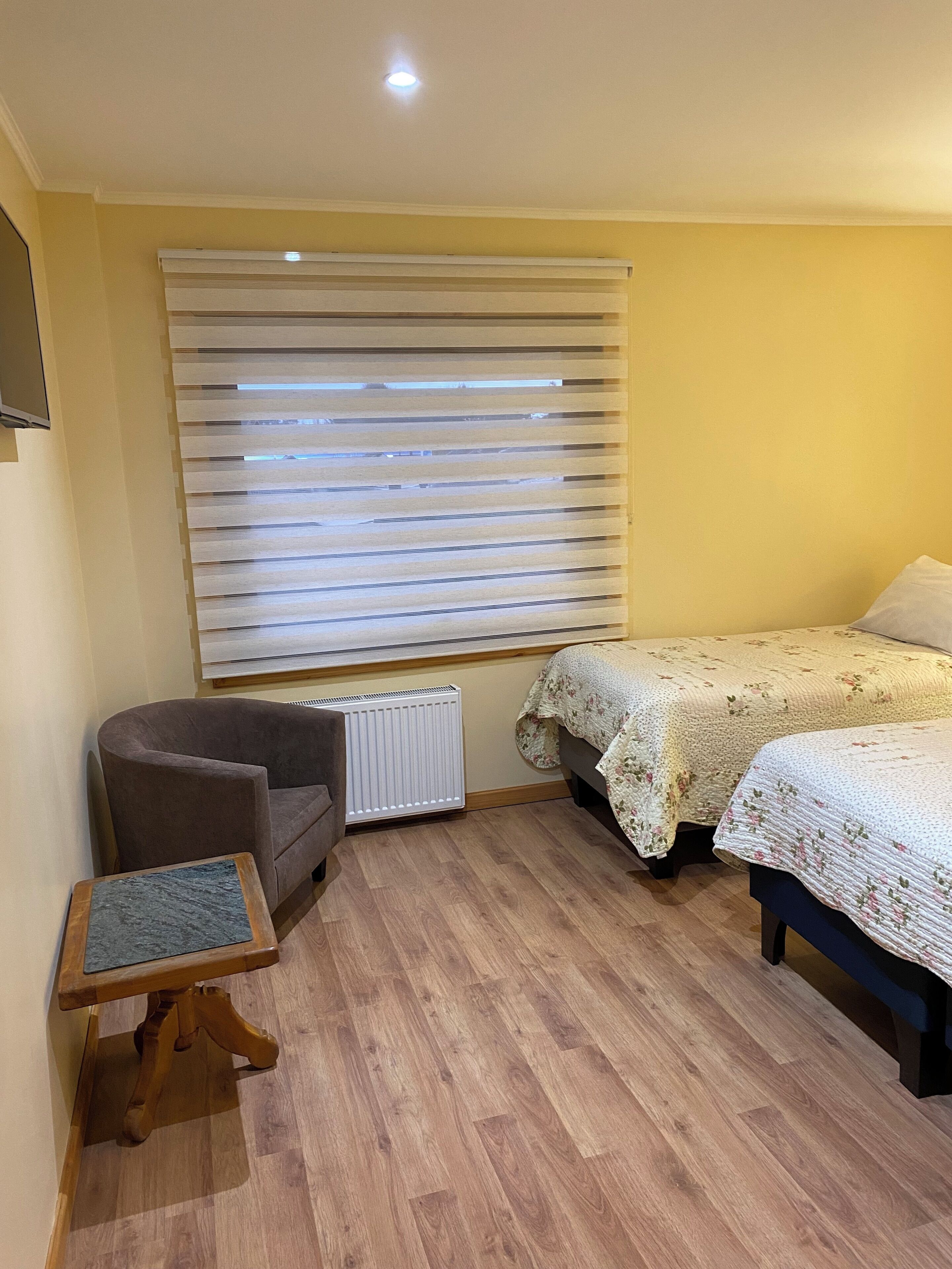 Un dormitorio con cama, una silla, una mesita, un radiador y una ventana con persianas.
