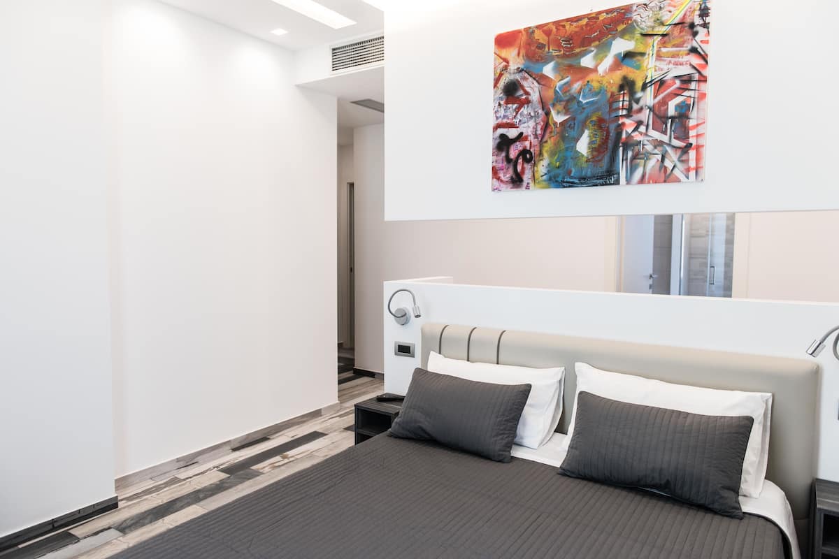 A modern hotel room with a large bed, a bedside table, a nightstand, and a wall-mounted artwork.