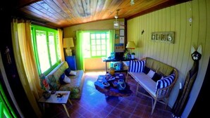 A cozy room with a wooden ceiling, a sofa, a small table, and a chair. There is a window with curtains and a lamp on a table. - Pousada Rosa da Praia (Imbituba)