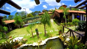 A tropical garden with a pond, palm trees, and a pathway leading to a building with a red roof. - Pousada Rosa da Praia (Imbituba)