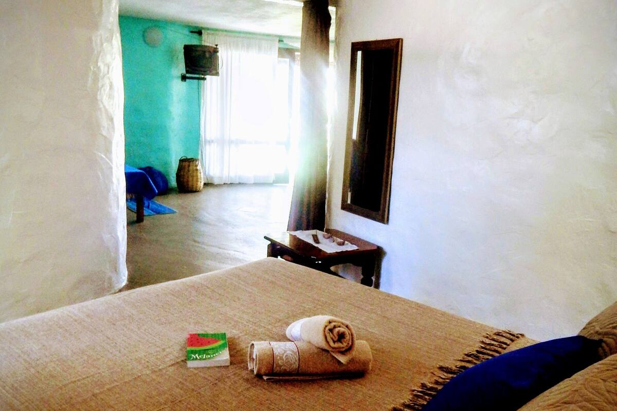 A hotel room with a bed, a small table, a mirror, and a view of the outside.