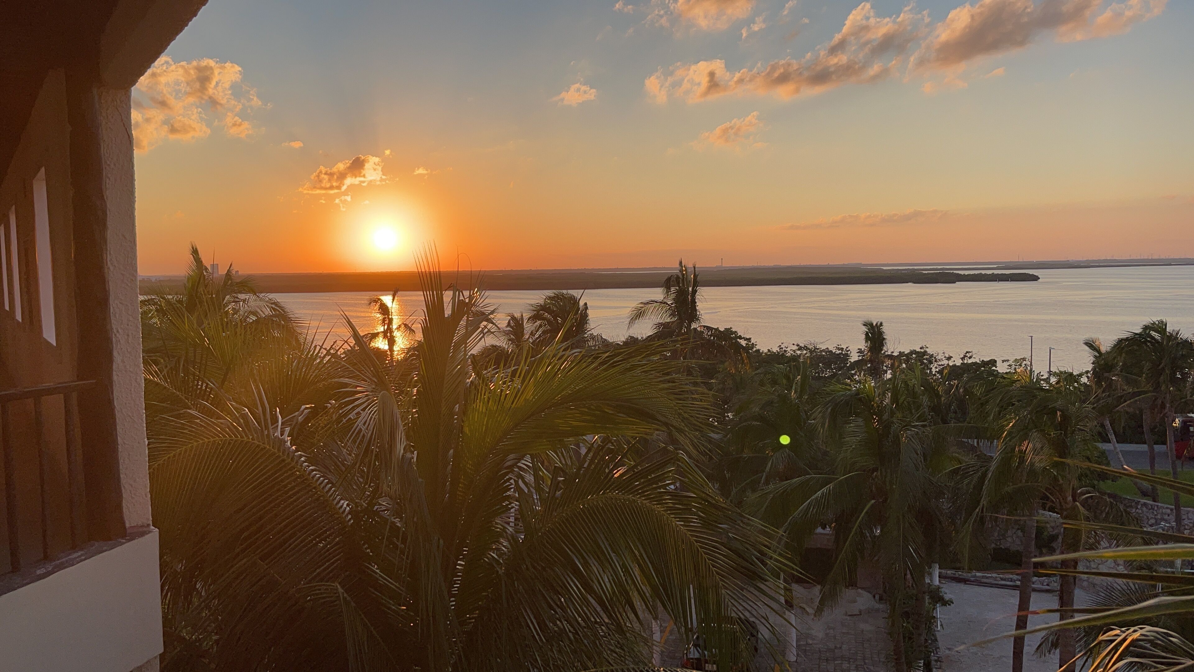 Ein Sonnenuntergangsblick von einem Balkon mit Blick auf ein Gewässer, Palmen und eine entfernte Küstenlinie.