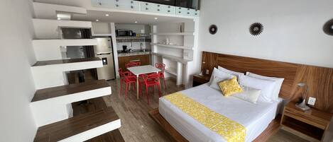Una habitación de hotel con cama, escritorio, silla, una mesita y una kitchenette.