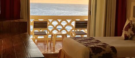 A hotel room with a balcony overlooking the sea, a bed with floral bedding, and a wooden desk.