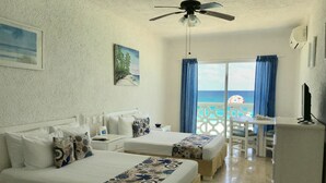A hotel room with two beds, a ceiling fan, a TV, a desk, and a view of the ocean. - Ocean View Apartments – Delicia (Cancun)