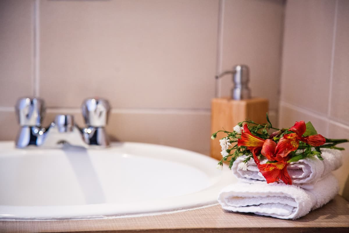Un baño con un lavamanos blanco, un jabonera de madera y una pila de toallas blancas con flores rojas.