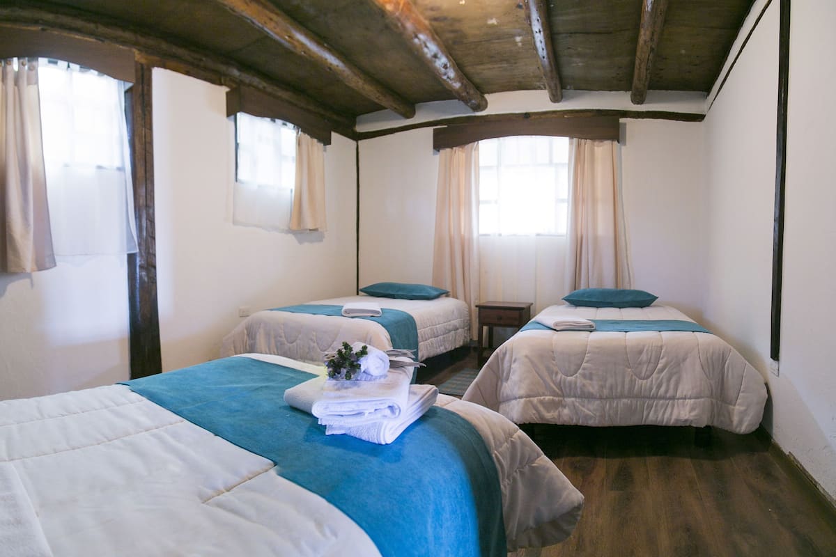 Una habitación con dos camas, cada una con una colcha azul y una almohada blanca. Hay ventanas con cortinas, vigas de madera en el techo y piso de madera.