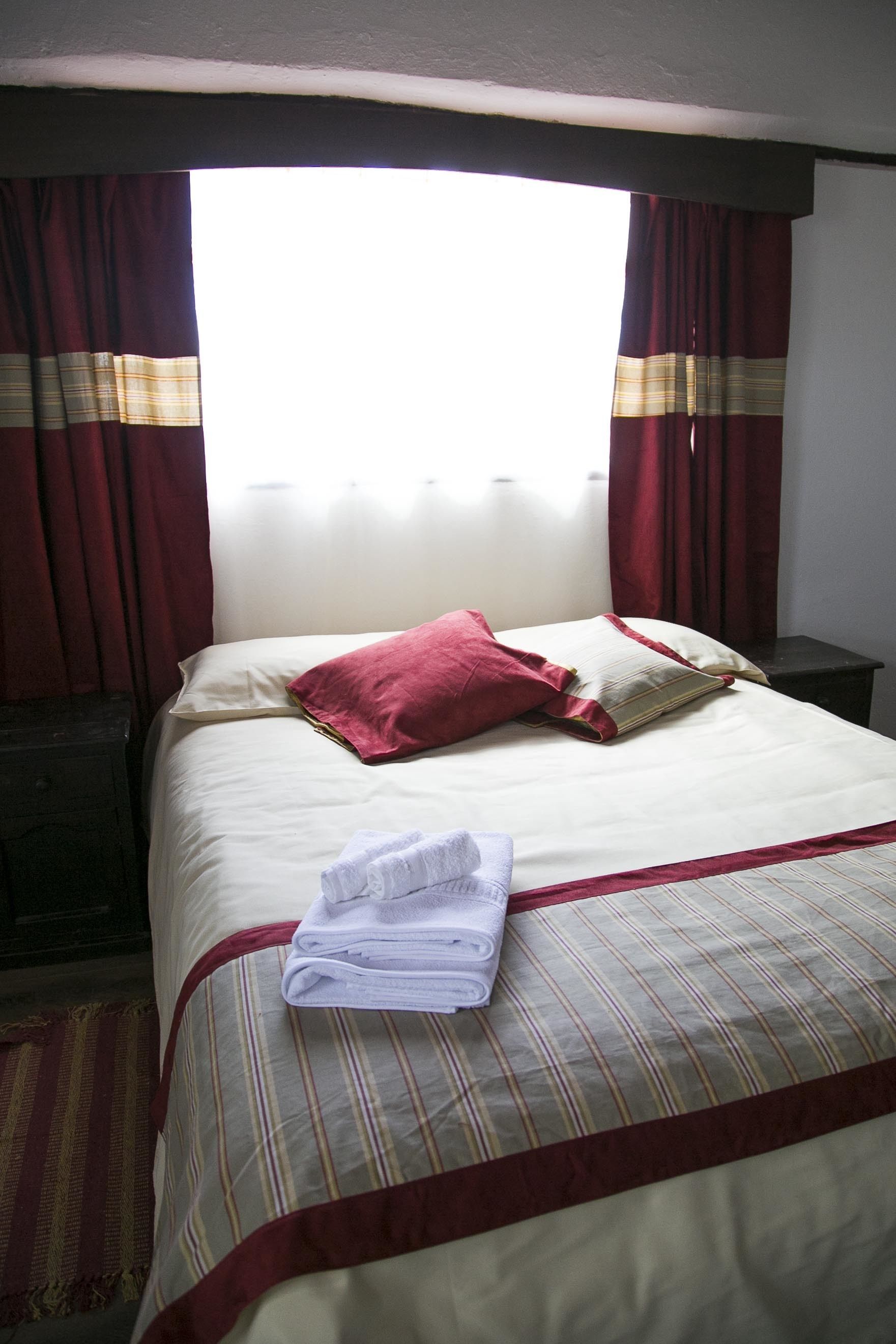 Una cama bien tendida con una colcha a rayas y dos almohadas, una toalla doblada sobre la cama, y una ventana con cortinas.