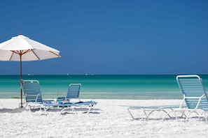 Una spiaggia con sabbia bianca, acqua cristallina turchese e un ombrellone bianco che offre ombra. Si vedono diversi lettini vuoti e un tavolino con un asciugamano.