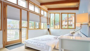 Une chambre à coucher avec un lit, une table de chevet et une vue sur un chalet de montagne, visible à travers de grandes fenêtres.