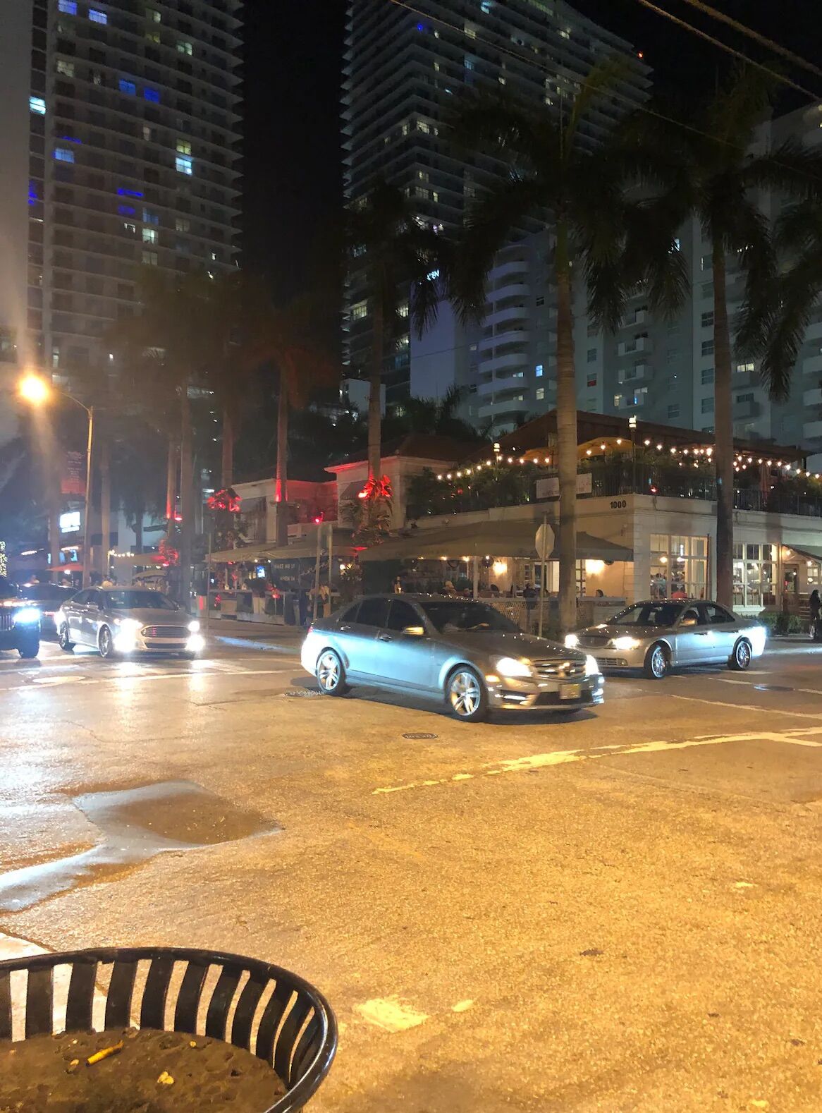 Calle iluminada de noche con coches, palmeras y edificios altos.