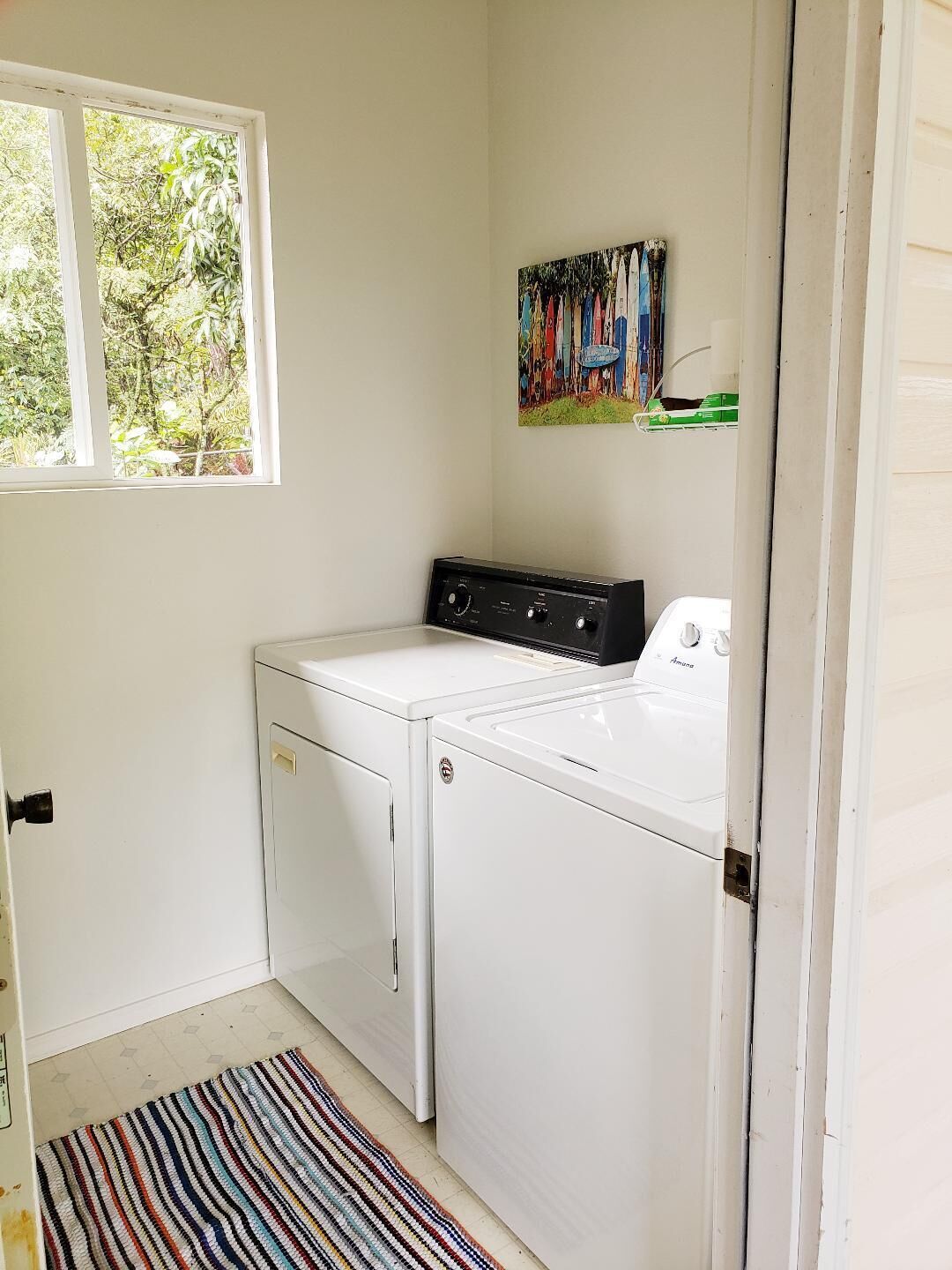 A laundry room with a washer and dryer, a window with a view of greenery, and a colorful painting on the wall.