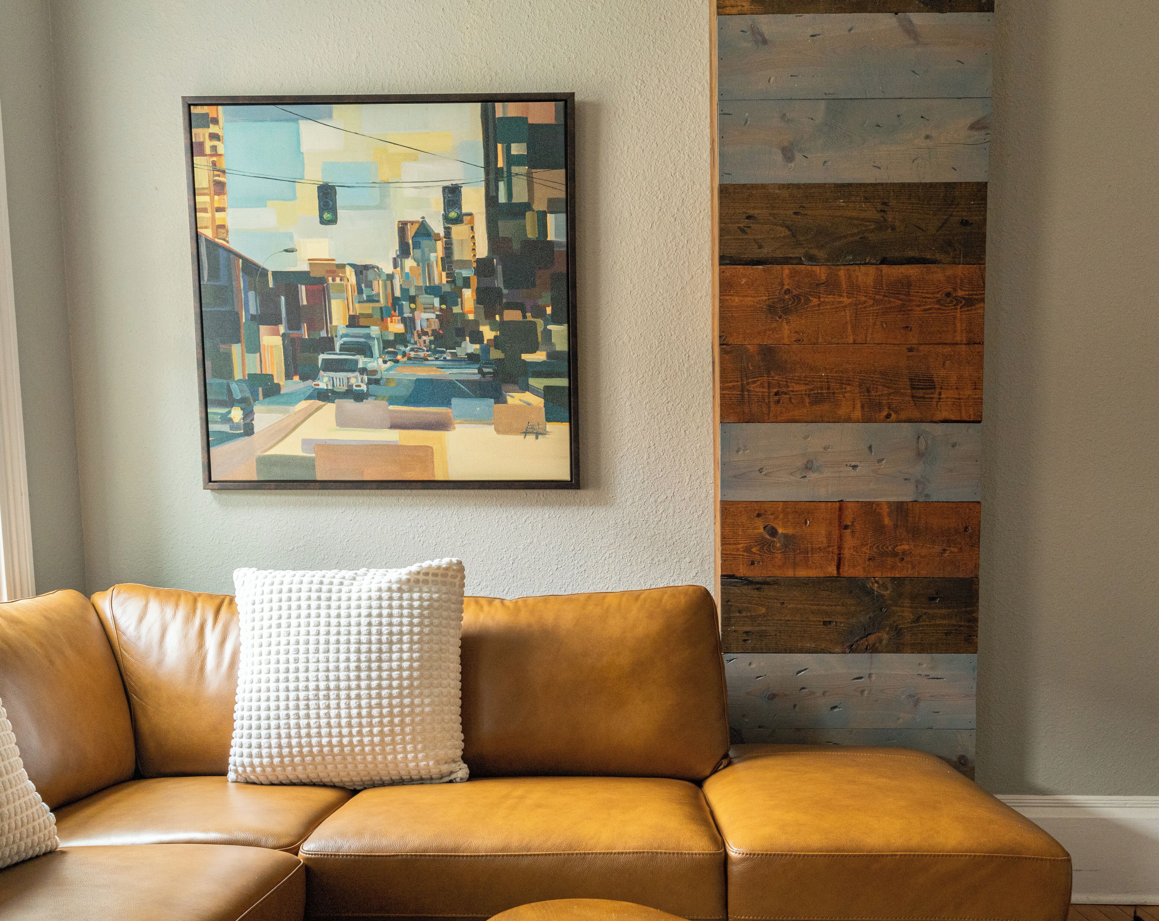 A brown leather sofa with a textured white pillow against a wall with a cityscape painting and a wooden panel.