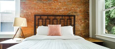 A bedroom with a brick wall, a wooden headboard, a white bedspread, a bedside lamp, and a window with a view of greenery.
