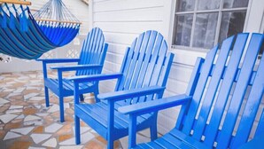 Three blue Adirondack chairs and a hammock on a tiled patio with a white wall and window in the background.
