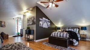 Une chambre avec un plafond en pente, un plancher en bois, un grand lit recouvert d’un plaid en fourrure, un bureau avec une chaise et une table de chevet avec une lampe.