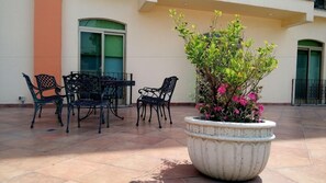 An outdoor seating area with a table and chairs, a large planter with flowers, and a building with a green door and windows. - Hg Hotel (Zapopan)
