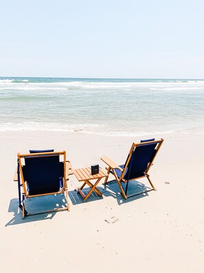 Zwei Holzliegen und ein kleiner Tisch an einem Sandstrand, im Hintergrund das Meer.