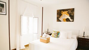 A neatly arranged bedroom with a bed, a bedside table, a nightstand, a window with curtains, and a painting on the wall.