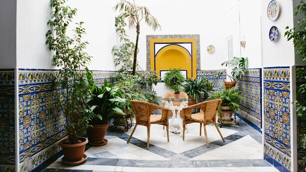 A courtyard with a tiled floor, potted plants, wicker chairs, and a central table.