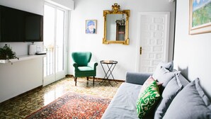 A living room with a green armchair, a grey sofa, a patterned rug, a large mirror, and a television on the wall.