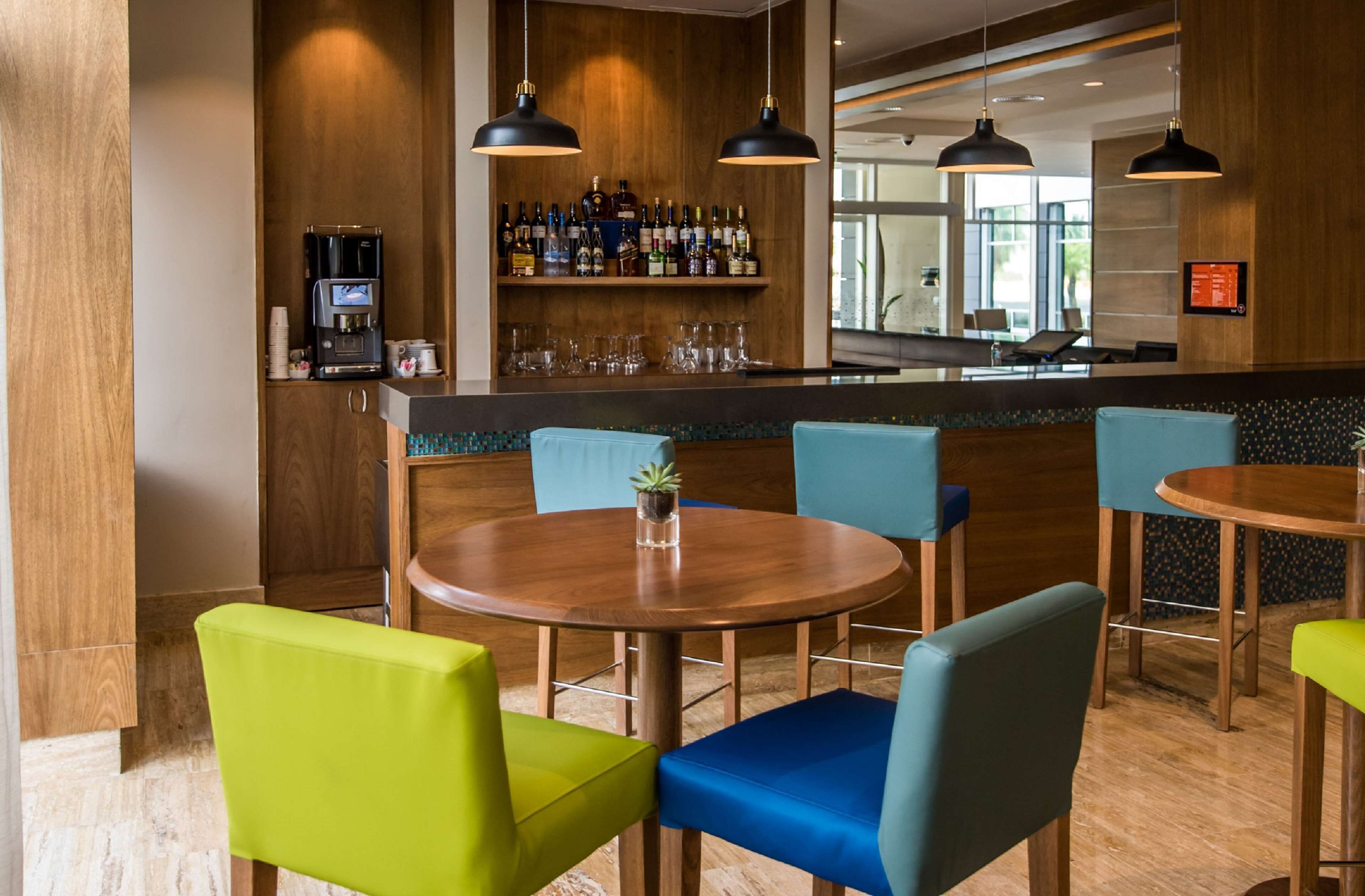 Zona bar moderna con pareti in legno, diverse bottiglie di liquori e una macchina per caffè. Sono presenti diversi tavoli e sedie con rivestimenti colorati.