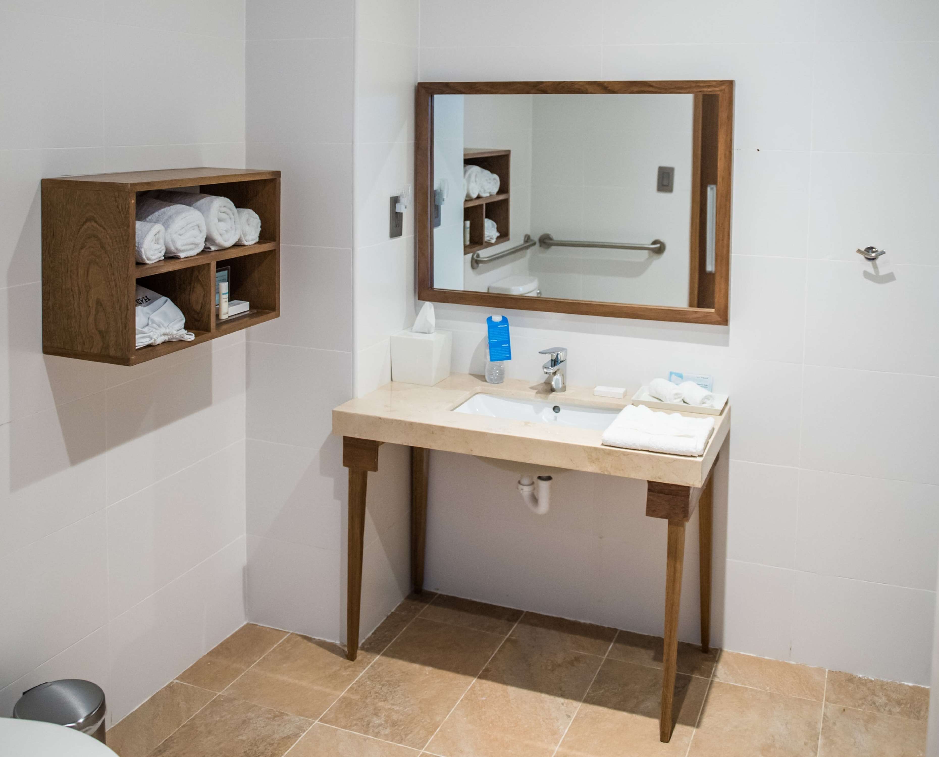 Un baño con lavamanos, espejo, toallero y una repisa de madera con toallas.