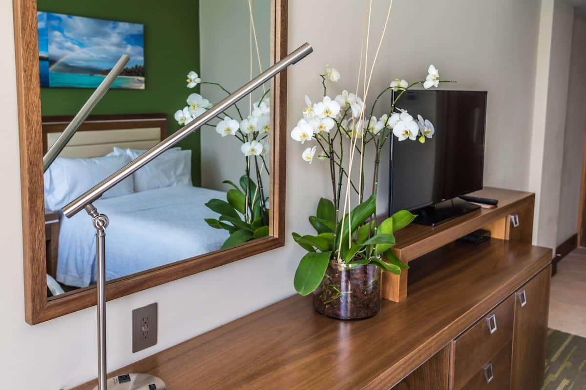 Una habitación de hotel con una cama, un televisor sobre un aparador de madera y orquídeas blancas en el aparador.