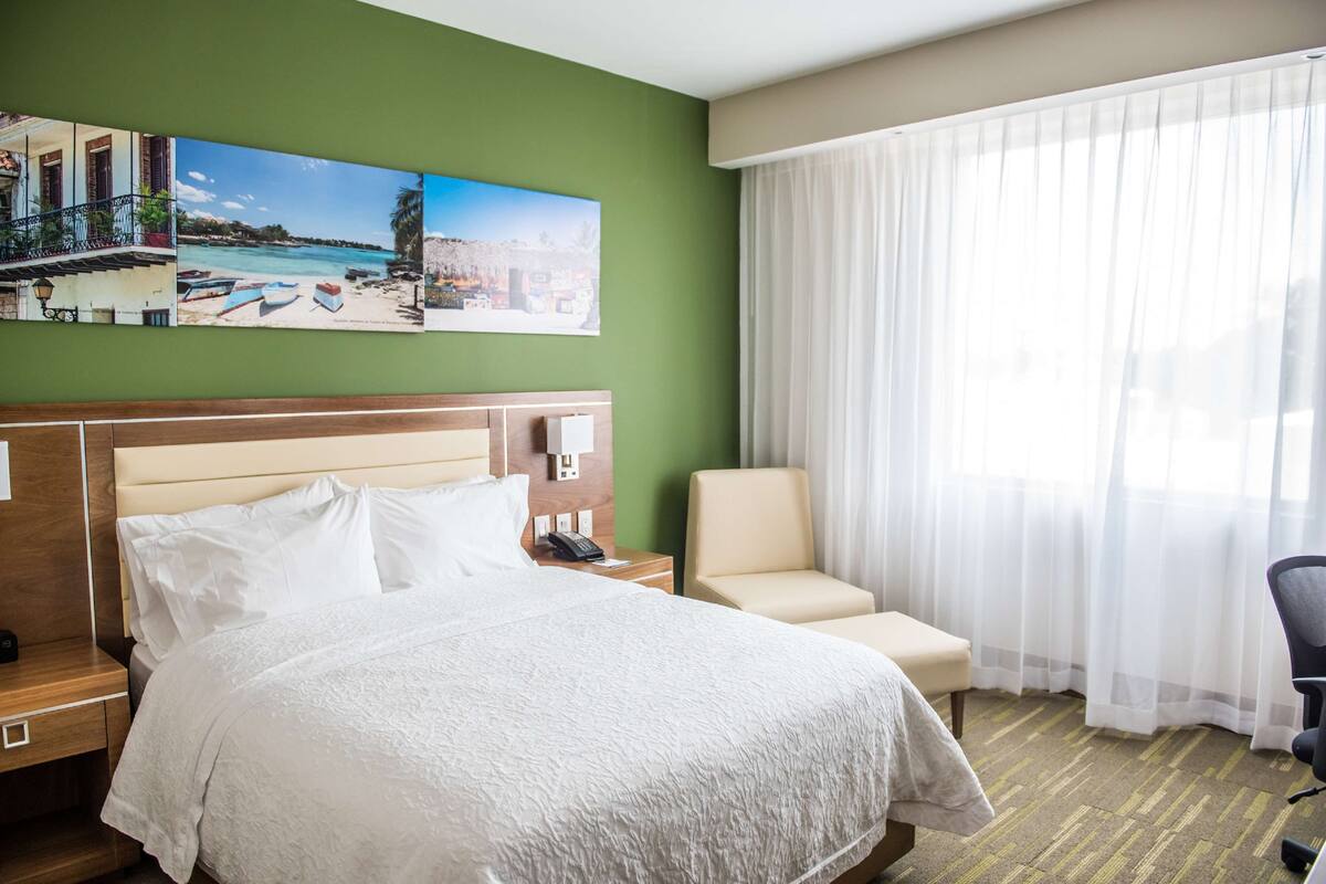 Habitación de hotel con una cama grande, una silla, una mesita de noche, un cuadro de una playa y una ventana con cortinas translúcidas.