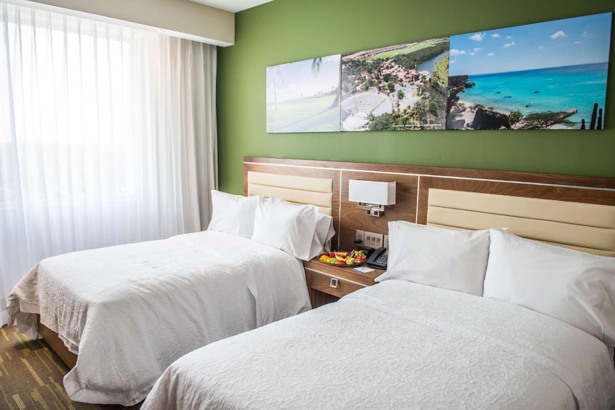 Habitación de hotel con dos camas, una mesita de noche con un teléfono y un frutero, y una pared con tres cuadros de paisajes.
