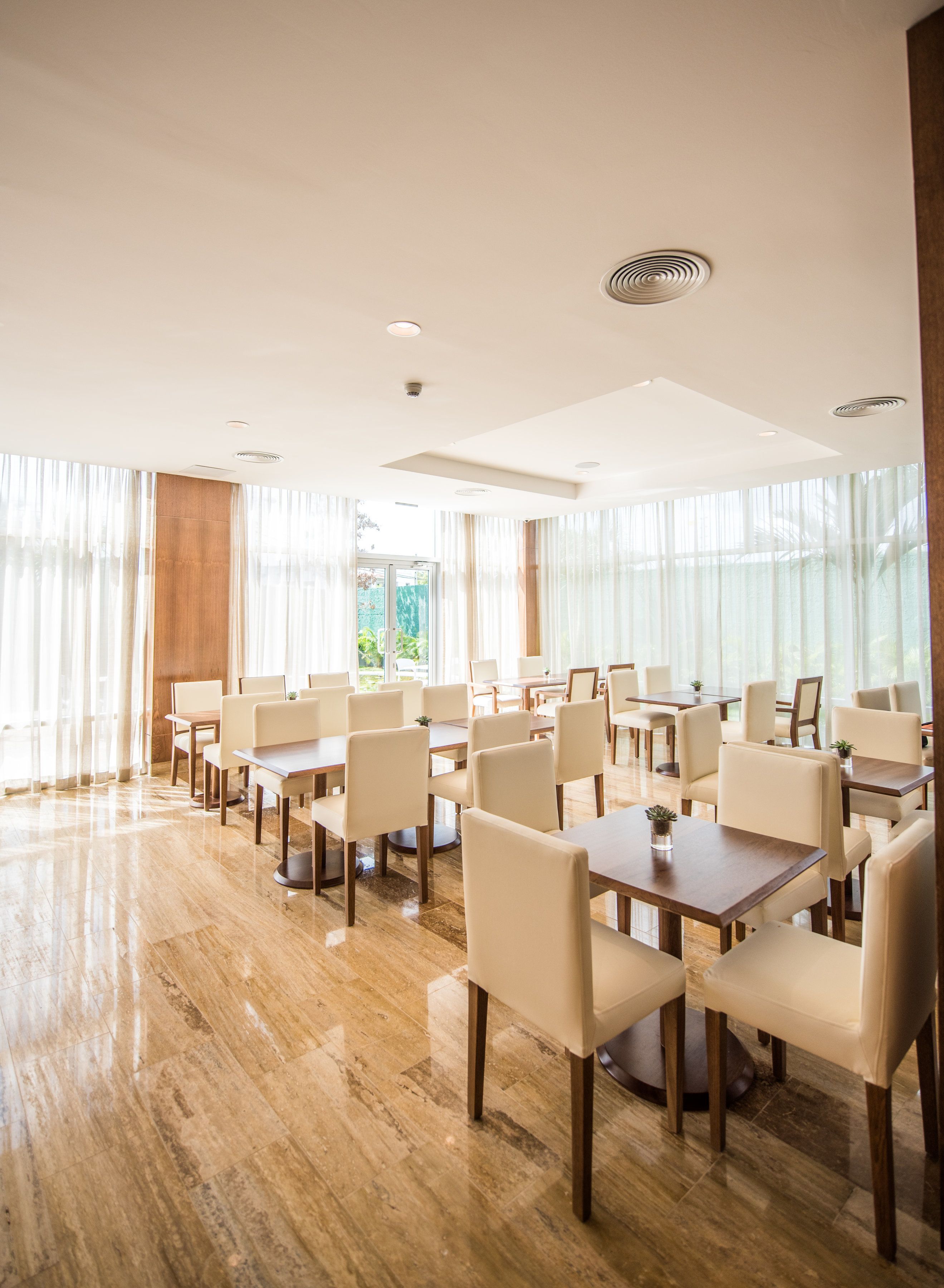 A spacious dining area with multiple tables and chairs, large windows with sheer curtains, and a polished wooden floor.