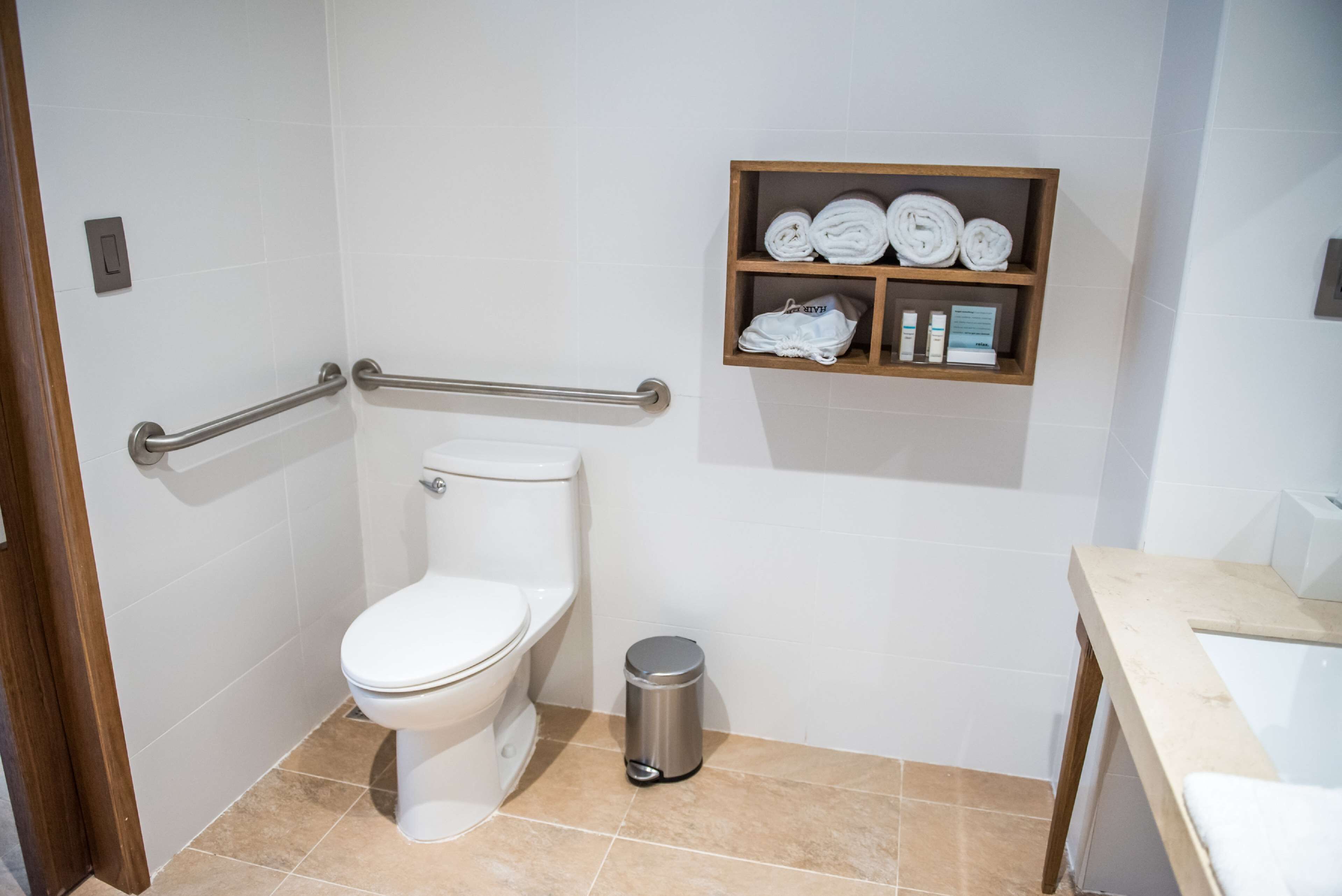 A bathroom with a toilet, a handrail, a shelf with towels and toiletries, and a trash bin.