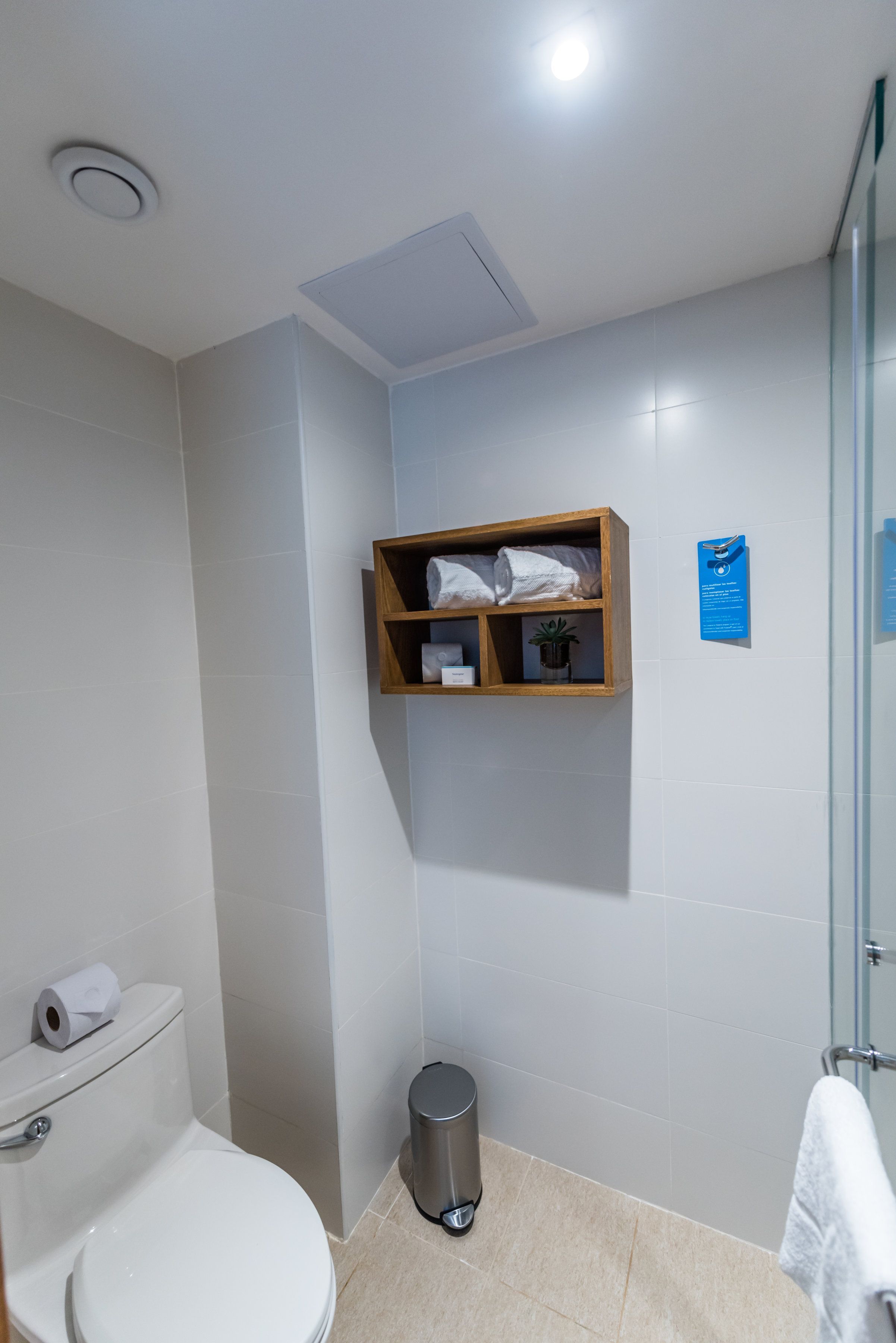 Un baño con inodoro, un cubo de basura, toallero, cabina de ducha de vidrio y estante de madera con artículos de tocador.