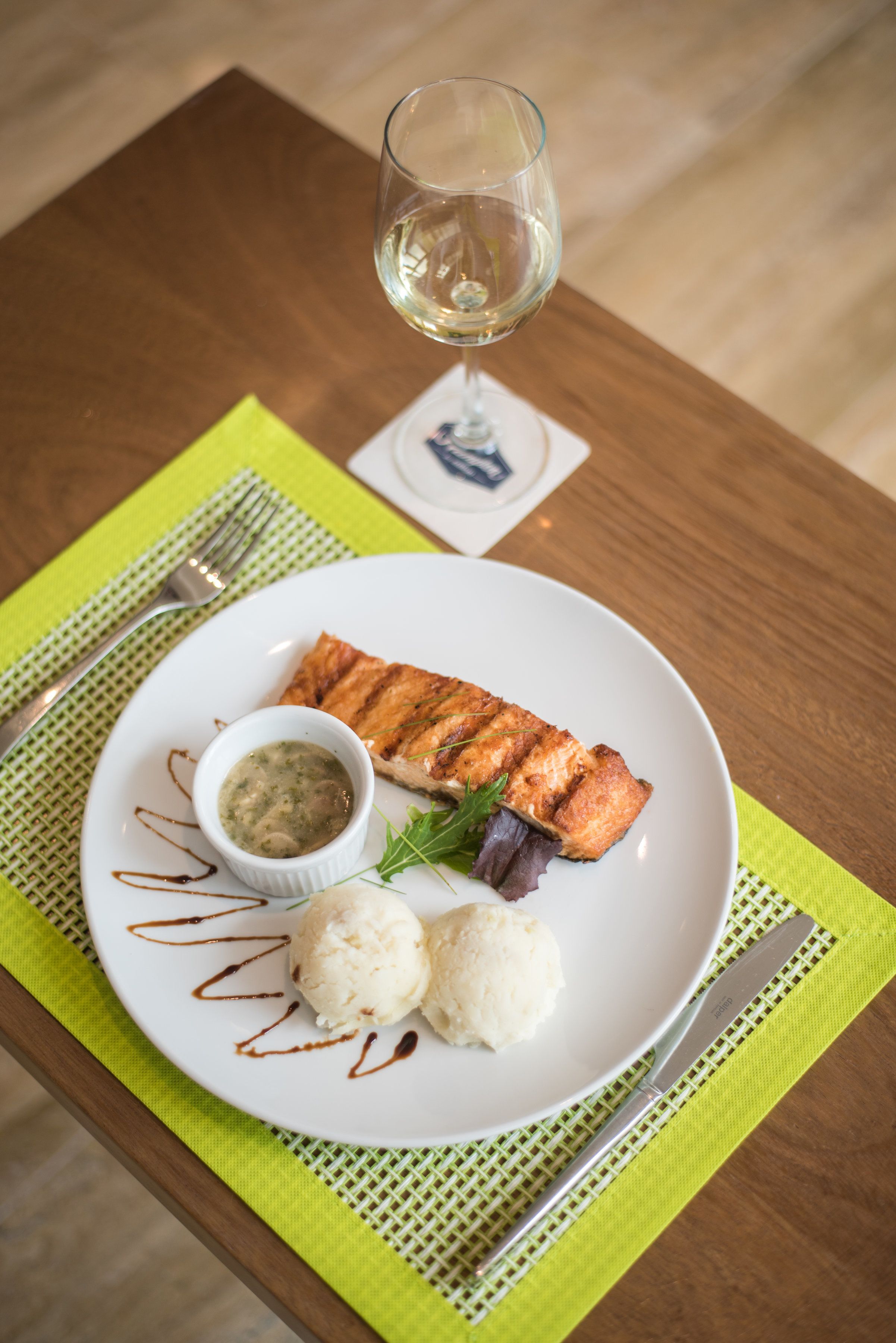 A plate with grilled salmon, a small bowl of sauce, and two scoops of ice cream on a white plate, placed on a green placemat.