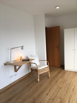 A room with a wooden floor, a white wall, a wicker chair with a white cushion, a small table with a lamp, and a white wardrobe. - Yellow House-Porto dos Carneiros Yellow House-Porto dos Carneiros (Lagoa, S. Miguel)