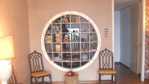 Une pièce avec un grand miroir rond au mur, deux chaises en bois avec des coussins, un petit panier en osier et une table avec une lampe.