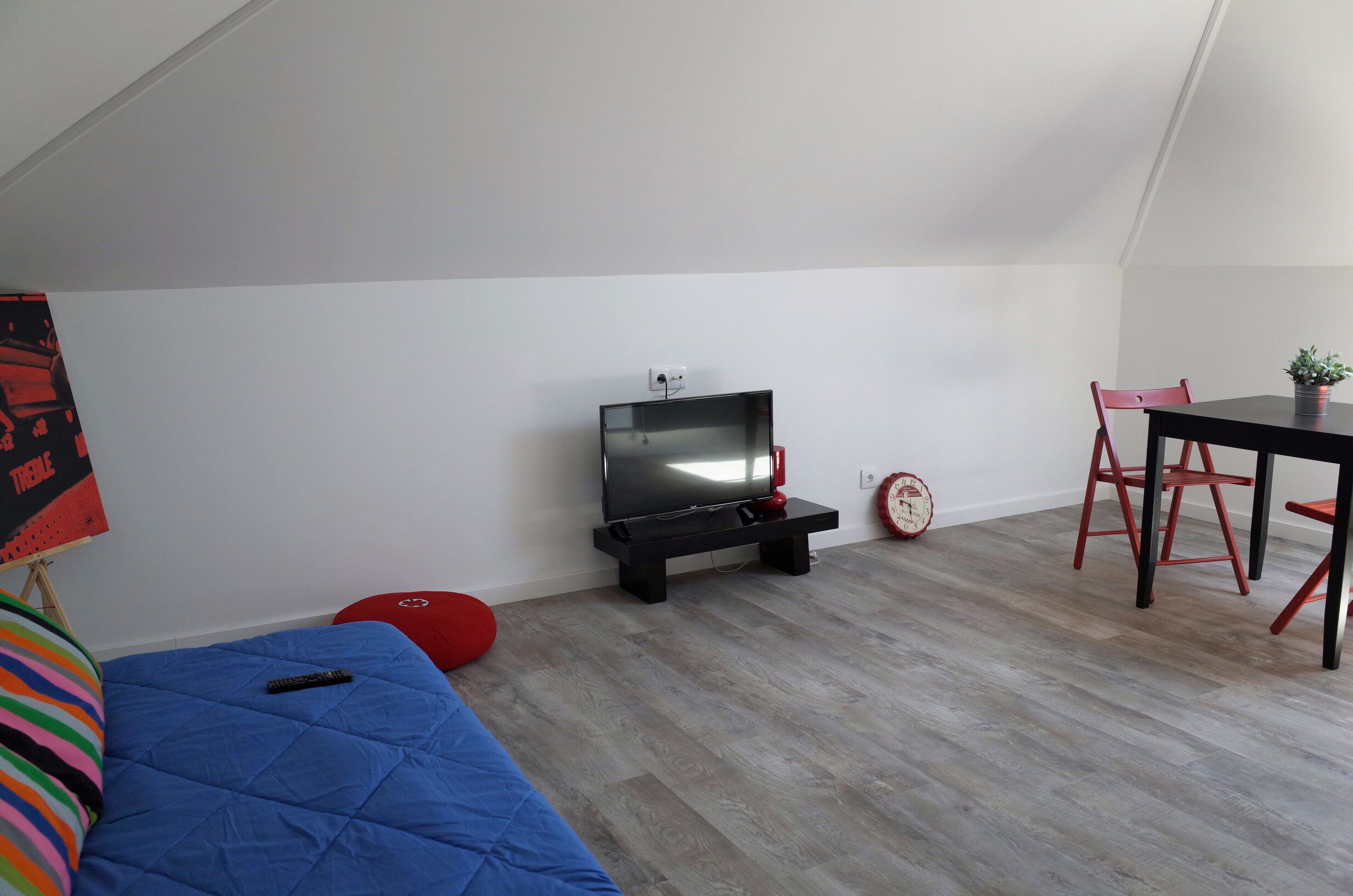 A small, cozy room with a TV on a stand, a dining table with chairs, a red bean bag, and a clock on the wall.