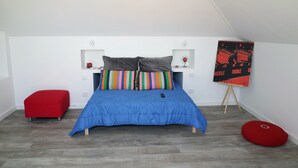 Een slaapkamer met een bed, een rode poef, een klein tafeltje met een vaas en een poster aan de muur.