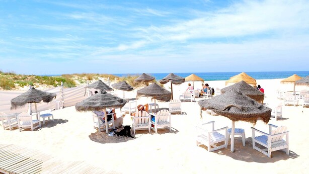 Een strand met rieten parasols en witte ligbedden.