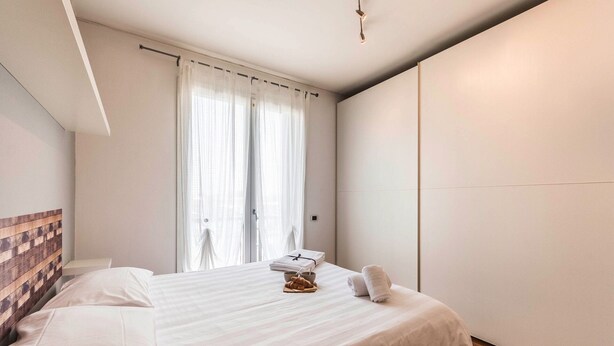 A hotel room with a bed, a wooden headboard, a window with sheer curtains, and a bedside table with a bowl and towels. - Novalesi Apartment (20 minutes from Venice) (Noale)