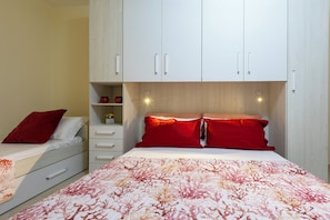 Um quarto com uma cama, dois travesseiros vermelhos, uma mesinha de cabeceira branca, uma cômoda e um guarda-roupa com um candeeiro sobre a cama.