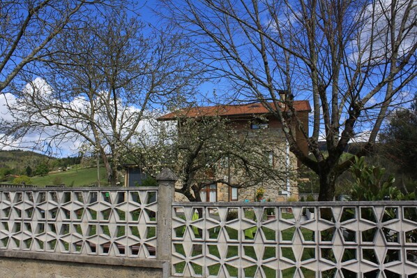 A stone house with a tiled roof, surrounded by trees and a decorative fence.
