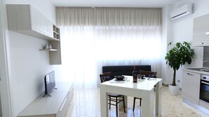 A modern kitchen with a dining area, white cabinetry, a large window with sheer curtains, and a potted plant.