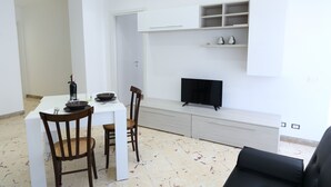 A modern living room with a dining table, two wooden chairs, a flat-screen TV mounted on a white wall, and built-in shelving above the TV.